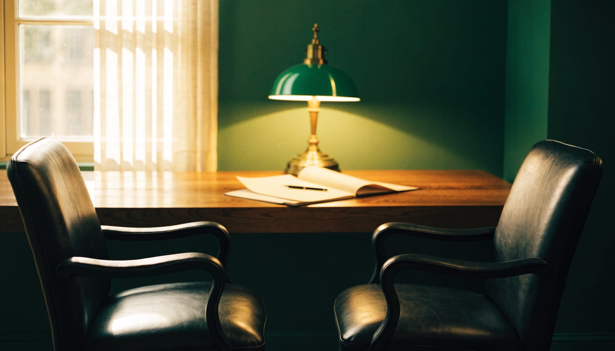Two leather chairs facing each other across a wide desk, a folder open between them under a green banker's lamp.