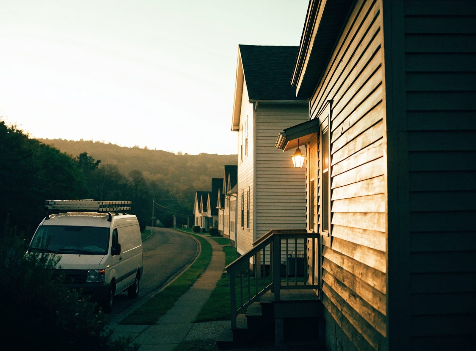 A contractor's van with ladders on its roof, parked before dawn on a street of wooden houses in the Hudson Valley.