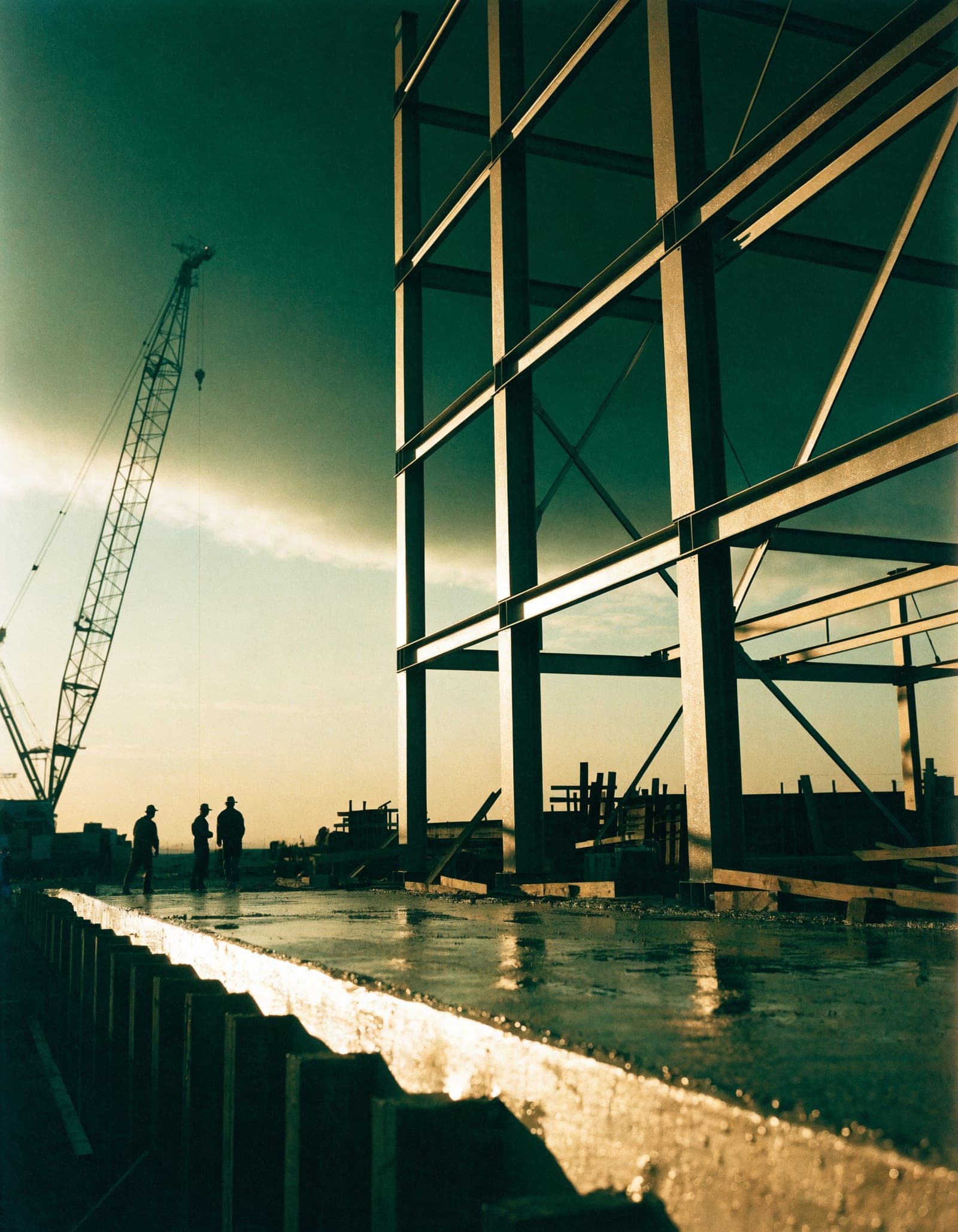 A construction site at first light: a steel frame rising, a crane in silhouette and a fresh concrete pour.