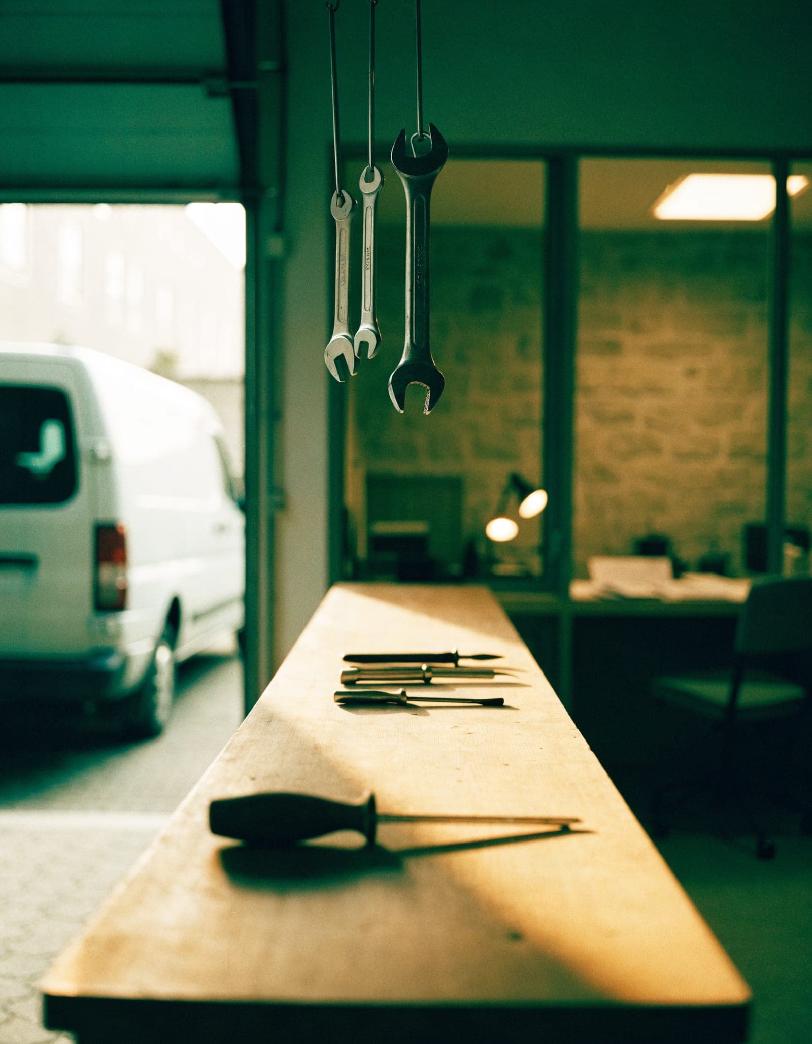 A small workshop at dawn: tools in a row on the bench, a work van in the open bay door, one light on in the back office.
