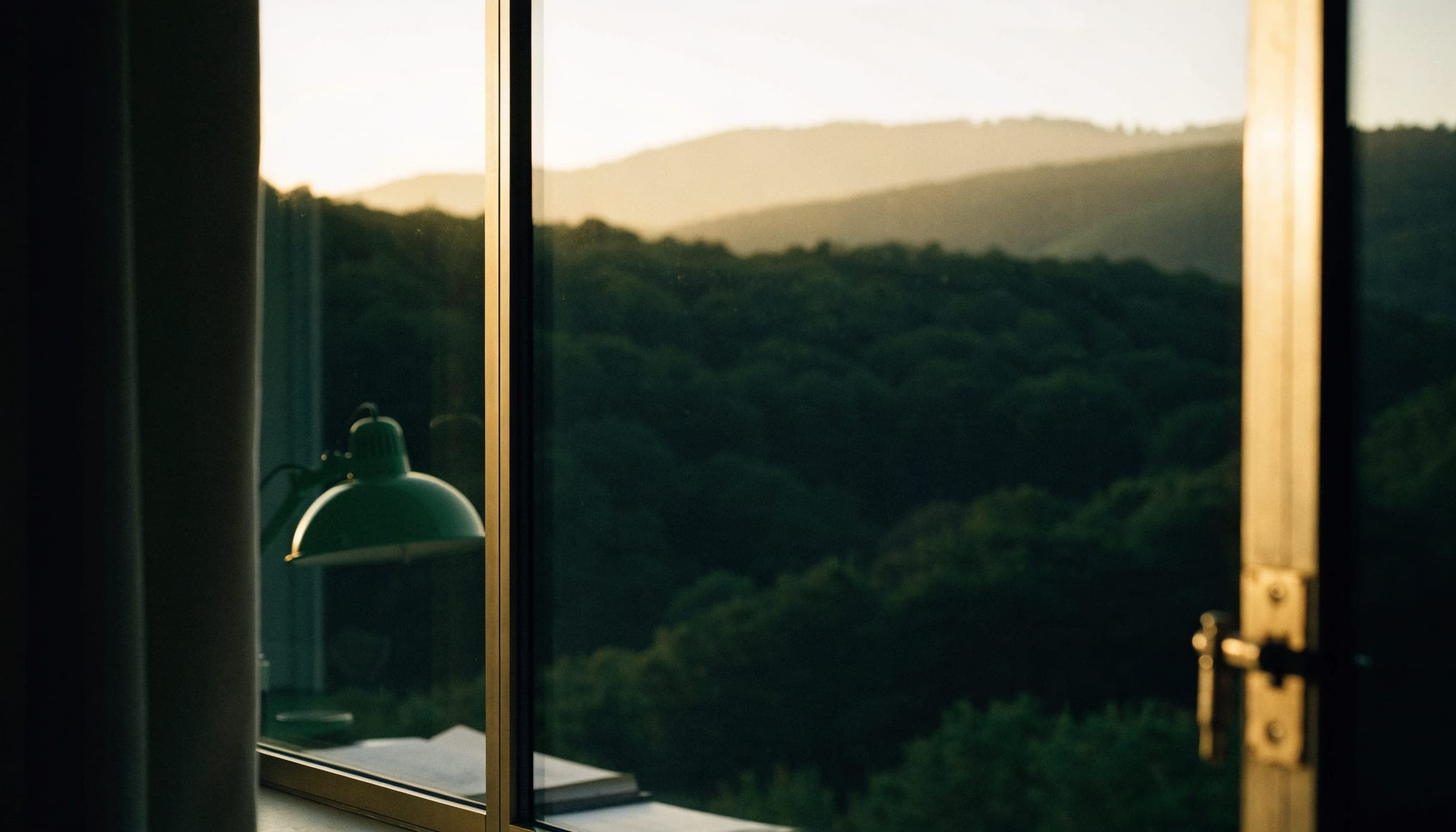 The view from an office window at dusk over the wooded hills of the Hudson Valley, a desk lamp reflected in the glass.